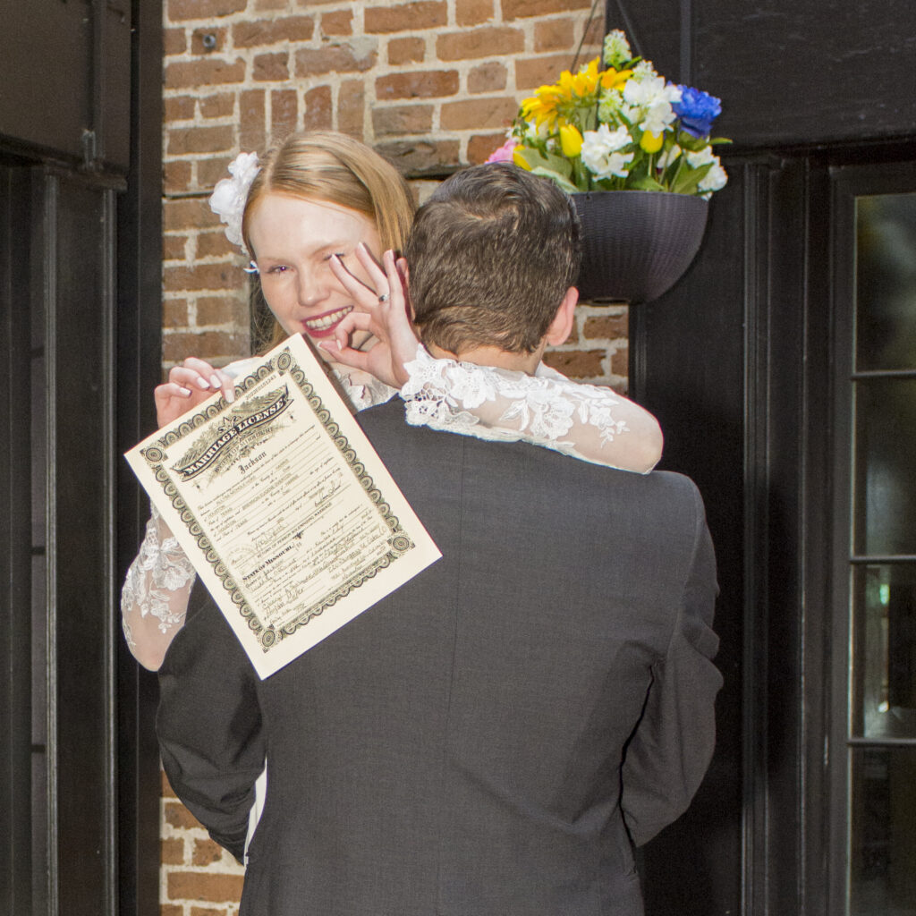 A bride being held my her groom with their wedding certificate after their special occasion